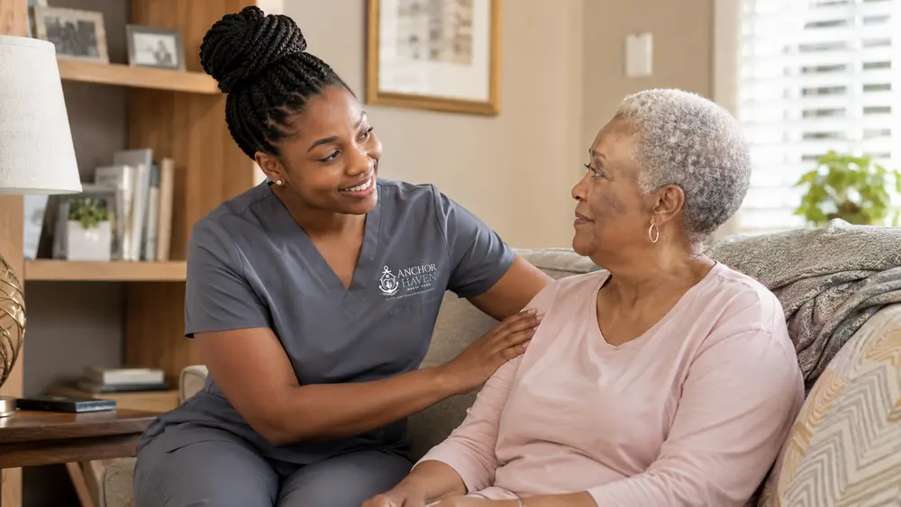 A caregiver with her hand resting gently on an older woman's shoulder as they talk on the sofa.